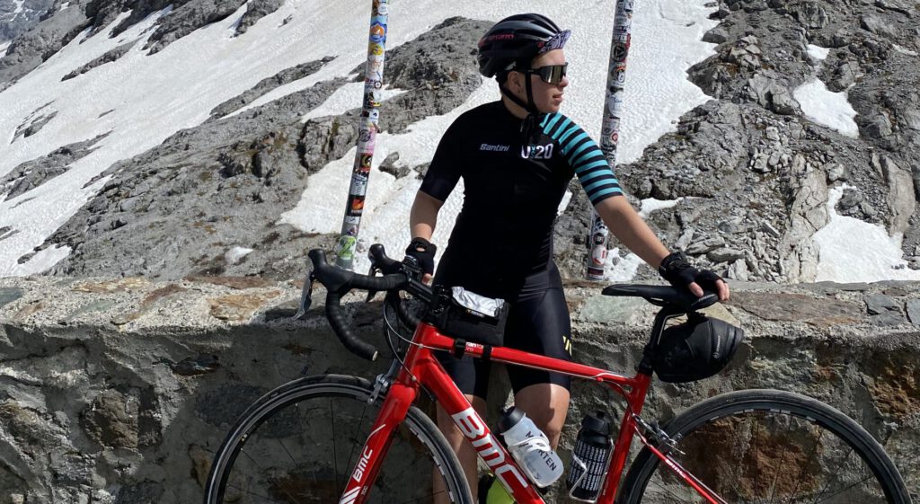Person im schwarzen Radtrikot mit rotem BMC-Rennrad vor Felsen und Schneeresten auf einem Alpenpass