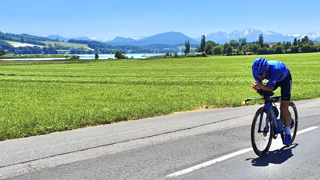 Triathlet in Aeroposition auf der Radstrecke am Obertrumer See, dahinter Felder, See und schneebedeckte Alpen
