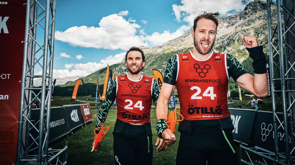 Swimrun-Duo mit Startnummer 24 jubelt beim Ötillö Engadin mit Paddles in den Händen vor Bergkulisse