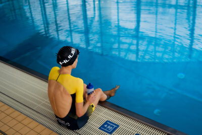 Triathletin im gelben Einteiler sitzt mit Trinkflasche am Beckenrand der Schwimmhalle bei den Arena Games