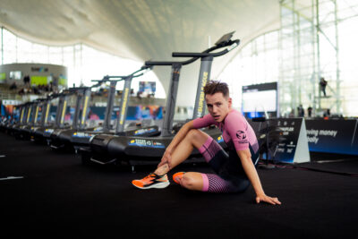 Triathlet im pinken Einteiler sitzt bei den Arena Games in München vor einer Reihe Laufbänder auf dem Boden