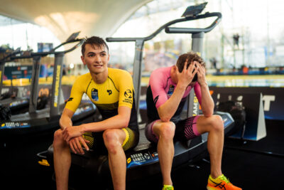Zwei Triathleten sitzen nach dem Rennen auf einem Laufband der Arena Games, einer lächelt, einer stützt den Kopf in die Hände