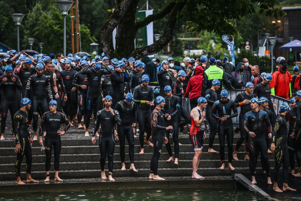 Triathleten in Neoprenanzügen und blauen Badekappen warten auf Stufen am Wasser auf den Schwimmstart