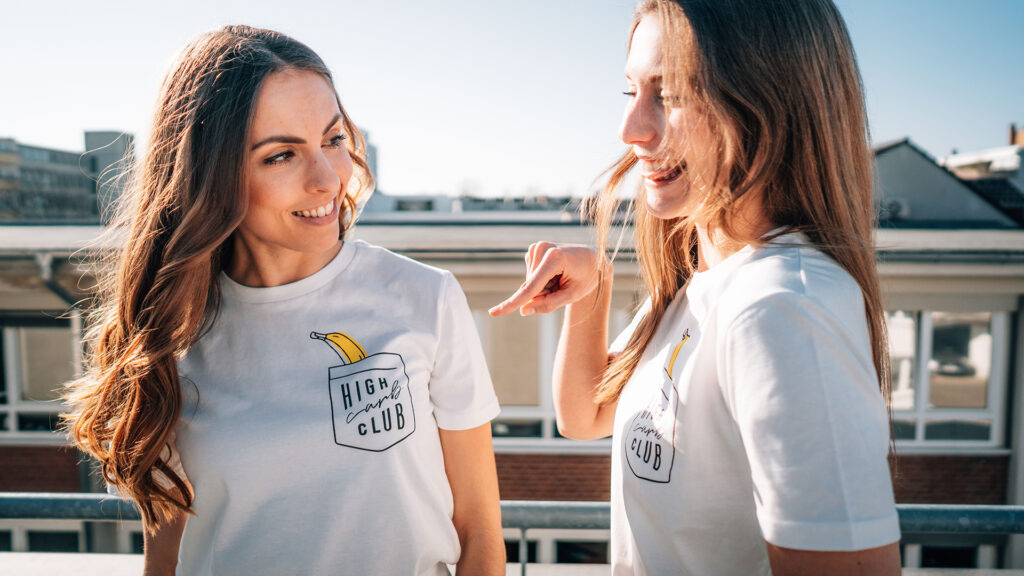 Zwei lachende Frauen in weißen T-Shirts mit High-Carb-Club-Aufdruck und Banane auf einer Dachterrasse