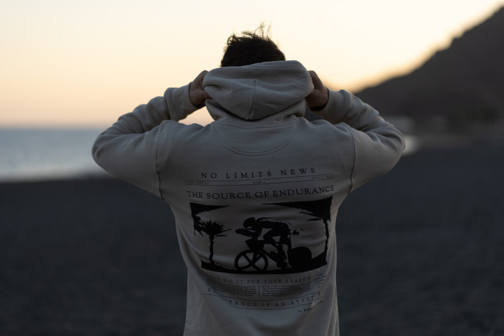 Person von hinten im Hoodie mit Aufdruck No Limits News – The Source of Endurance an einem dunklen Strand