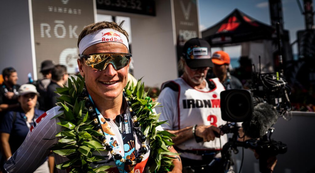 Lachender Triathlet mit Stirnband und Blumenkranz im Ziel der Ironman-WM auf Hawaii, dahinter ein Kameramann