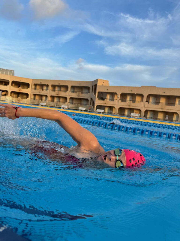 Schwimmerin mit pinker Badekappe krault im Außenbecken vor einer Hotelanlage