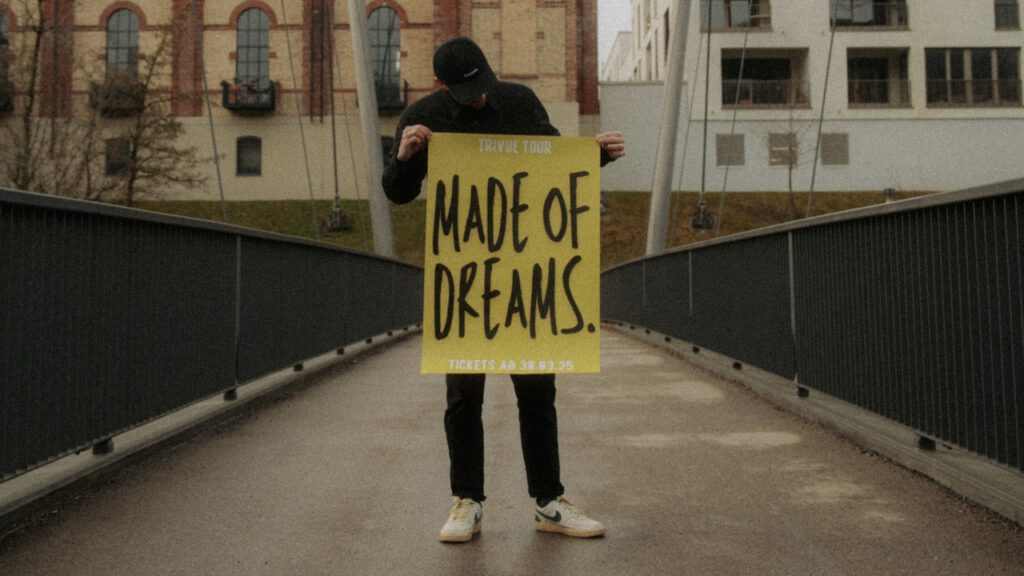 Person hält auf einer Fußgängerbrücke ein gelbes Plakat der Trivue Tour mit der Aufschrift Made of Dreams