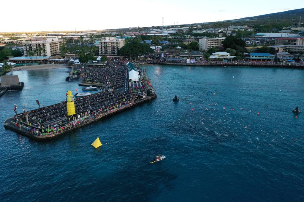 Luftaufnahme vom Pier in Kailua-Kona beim Schwimmstart: Wechselzone voller Räder, Zuschauer am Ufer, Schwimmerinnen und Schwimmer in der Bucht