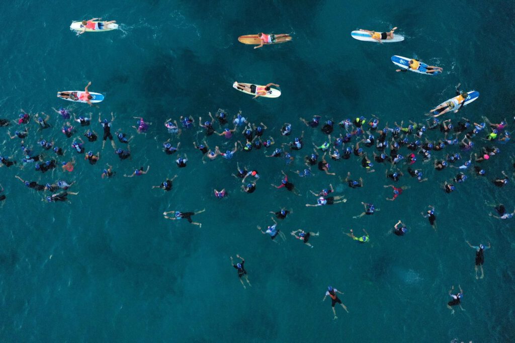 Luftaufnahme: Dutzende Schwimmerinnen und Schwimmer mit blauen Badekappen im türkisen Wasser, davor Helferinnen und Helfer auf Surfbrettern